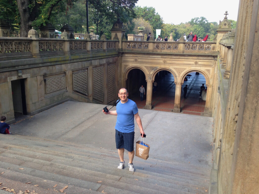 Michael standing at the top of steps leading down to Bethesda fountain in Central Park.He has a blue t shirt and shorts on and is carrying a bag and camera. The steps lead down to three archways and into a tunnel.